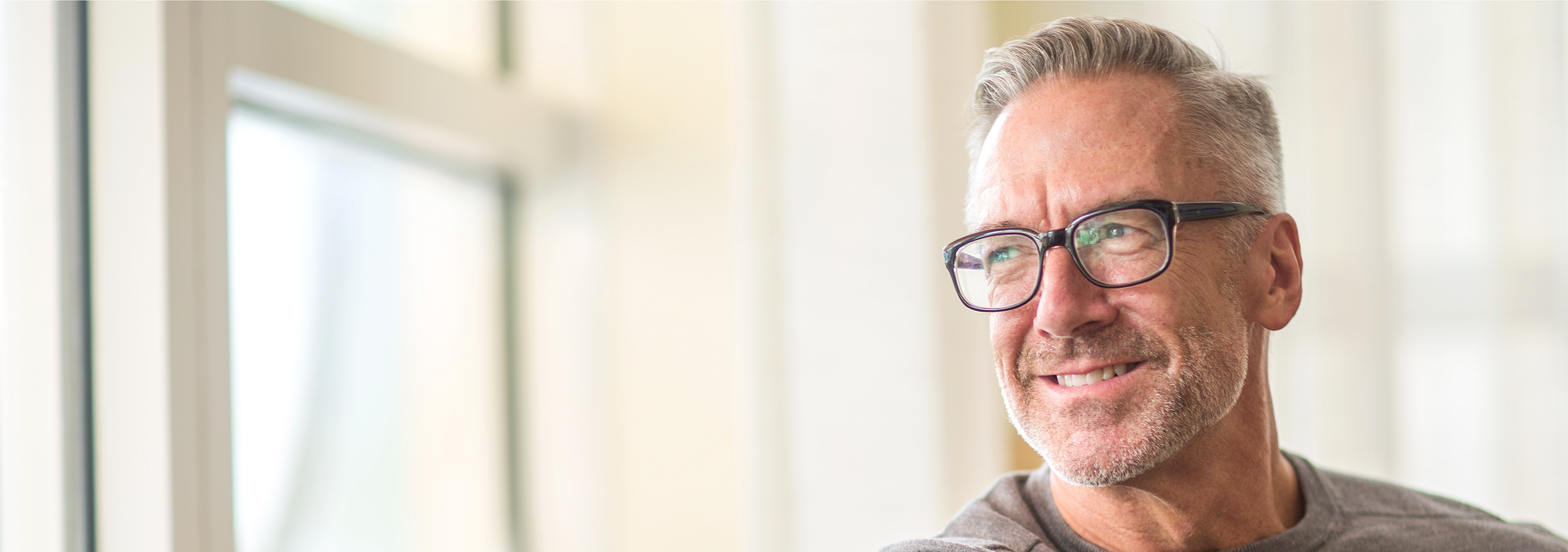 A male in his 50s wearing eye glasses and smiling while looking outside through a window.